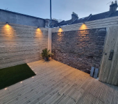 Modern garden patio with wooden decking, ambient wall lights, and a potted plant near a brick and wood-panelled fence
