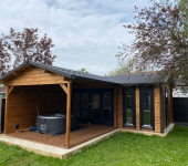 Wooden garden room with large windows and a hot tub on a patio under a cloudy sky surrounded by trees and grass