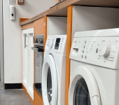 Row of modern white washing machines in a laundry room with orange accents and an exit door with blinds