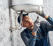 Electrician in plaid shirt and cap repairing a white water heater on a tiled wall using a screwdriver and gloves
