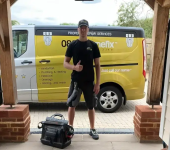 Handyman in black uniform posing with toolkit in front of yellow service van, offering repair services at a home entrance
