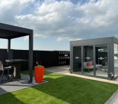 Modern garden with glass summerhouse, patio set, vibrant orange planter, and lush green lawn under a cloudy sky
