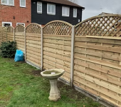 Wooden garden fence with trellis top, beside a house with lawn and birdbath, cloudy sky in the background