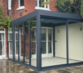 Modern glass conservatory extension on a brick house surrounded by greenery and a stone patio