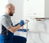 Technician inspecting boiler with clipboard, wearing blue overalls and gloves in a modern home setting
