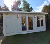 White garden shed with large windows and a Yale lock, surrounded by trees and a wooden fence on a sunny day
