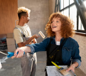 Two people painting a room with paint rollers, standing by large windows, wearing casual clothes and smiling