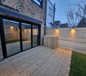 Modern wooden patio with glass doors and warm lighting, featuring a fenced yard and newly installed decking