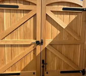 Wooden double gate with black metal hinges set against a blue sky and cobblestone driveway