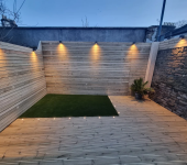 Modern wood-panelled garden with decked flooring and ambient lighting surrounding a small artificial grass area