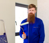 Man in blue overalls holding a paint roller beside a ladder indoors
