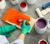 Person in green gloves painting wood with orange paint surrounded by open paint cans and brushes on a grey surface